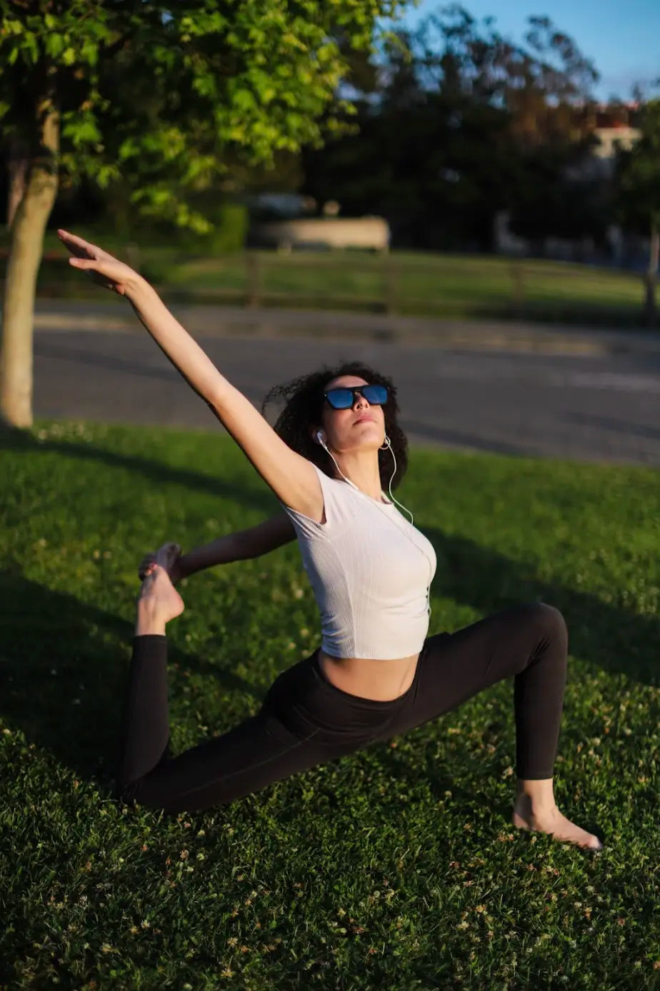 Woman stretching and doing yoga on the grass while wearing Healyan glasses and wired headphones